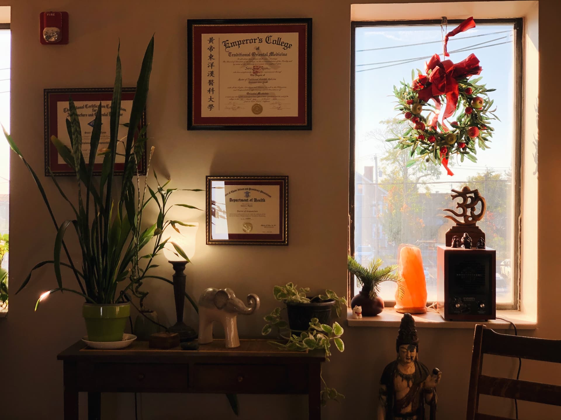 Framed diplomas beside a sunlit window with a wreath and Buddha statue